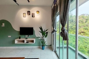 A living room with a TV, a plant, and a large window with curtains. - Icolor Home & Stay CH4 (Tanah Rata)
