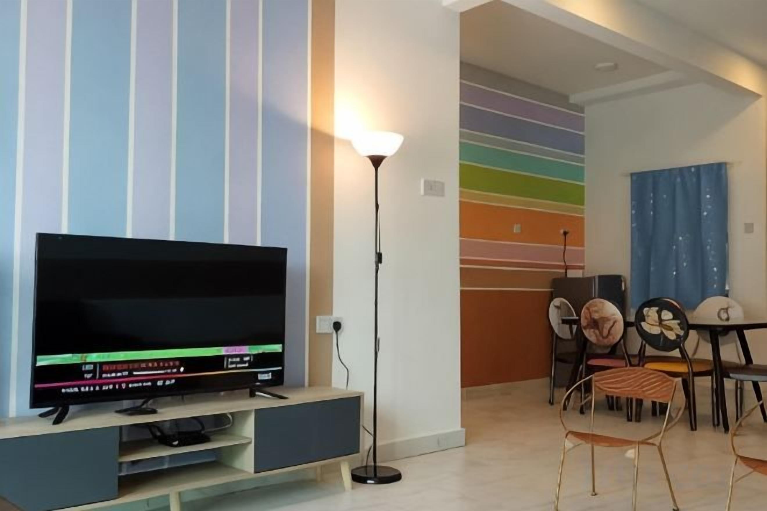 Una sala de estar moderna con un televisor de pantalla plana sobre un mueble, una lámpara de pie y un comedor con sillas y una mesa.