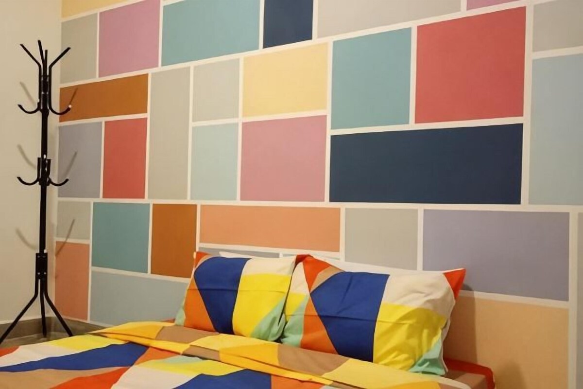 Un lit recouvert d’une couverture aux couleurs vives, un porte-manteau et un mur orné d’un motif géométrique.