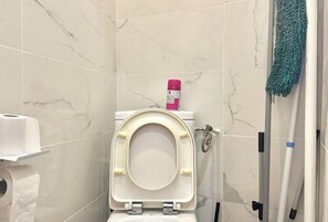 A bathroom with a toilet, a roll of toilet paper, a pink bottle, and cleaning tools. - Chic & Modern Studio in Centre Paris Rambuteau (Paris)