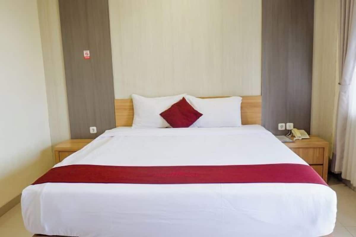 Una camera d'albergo con un letto grande, un comodino in legno con un telefono e un cuscino decorativo rosso.