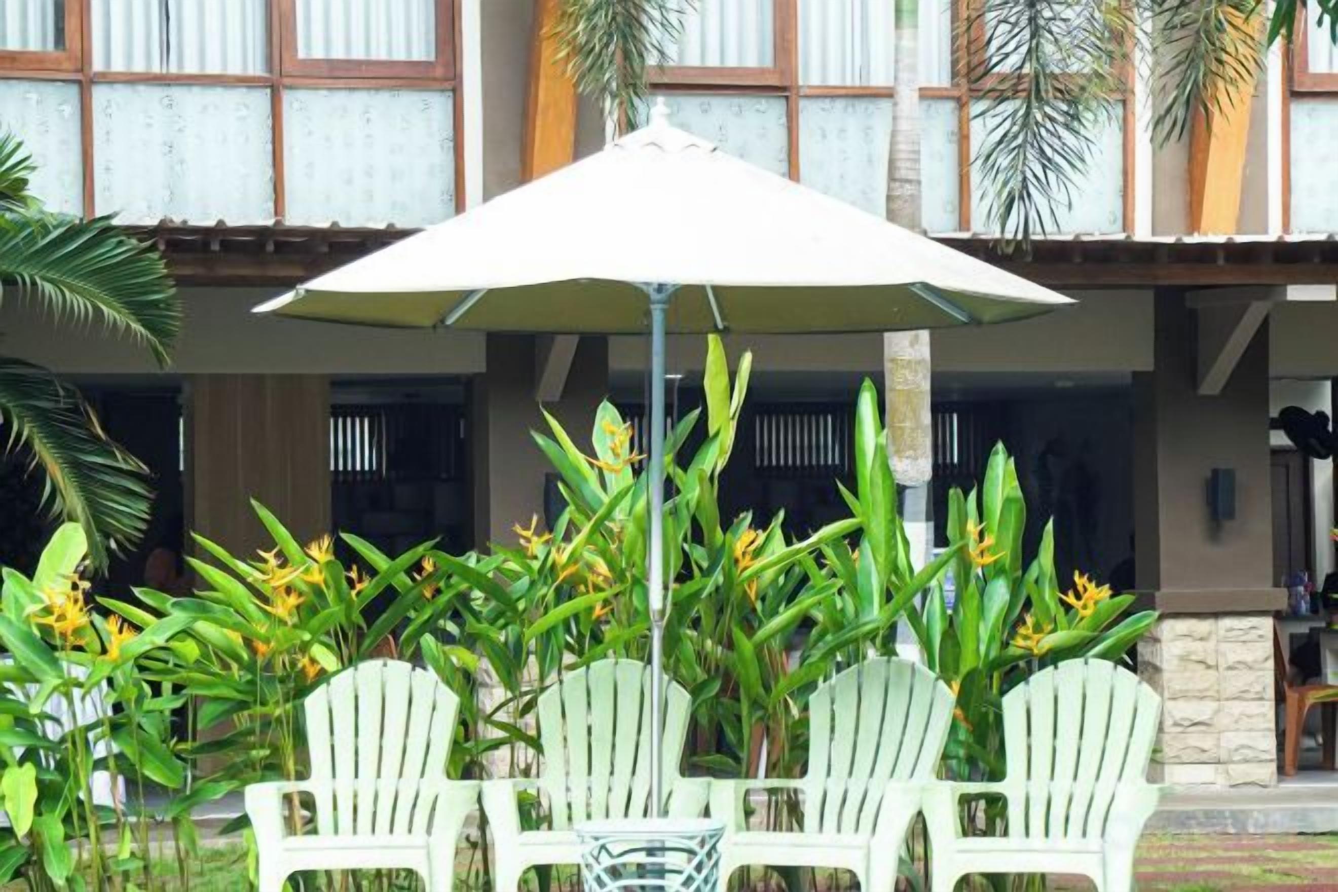 Een terras met drie witte stoelen en een tafel, beschut onder een grote parasol, omringd door tropische planten.
