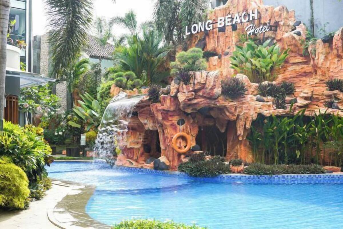 Area piscina con cascata, palme e formazione rocciosa con la scritta 'LONG BEACH Hotel'.