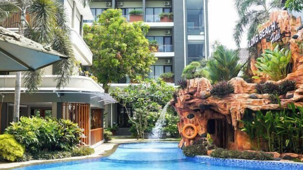 A hotel with a pool area featuring a rock formation with a waterfall, surrounded by lush greenery and a multi-story building in the background. - Long Beach Hotel Pangandaran (Pangandaran)
