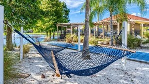 A hammock on a sandy beach with a pool and palm trees in the background.