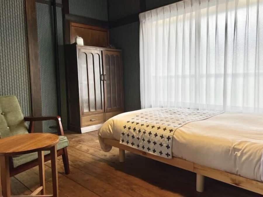 Un dormitorio con una cama de madera, un sillón verde, un armario de madera y una ventana con cortinas translúcidas.