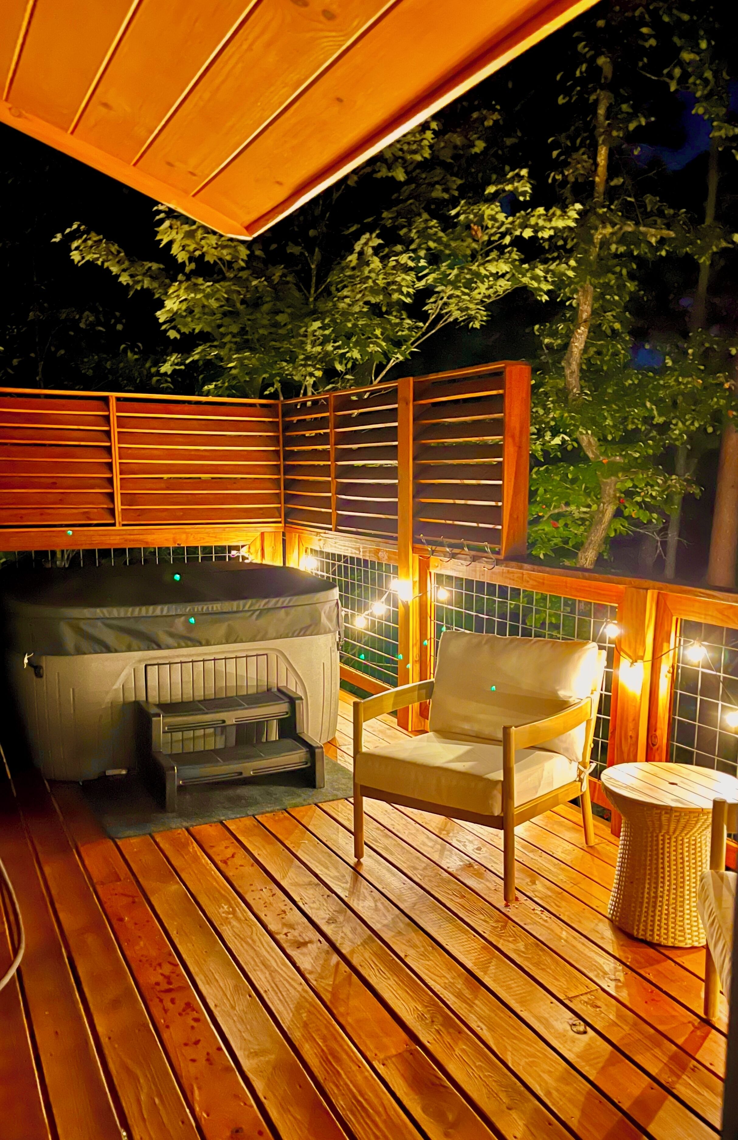 A wooden deck with a hot tub, a chair, and a table, surrounded by a wooden fence and trees.