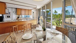 A modern kitchen with wooden cabinets, a dining area with a glass table, and a balcony with a view of palm trees and a distant mountain. - Kamaole Nalu 506 - Ocean Front (Kihei)