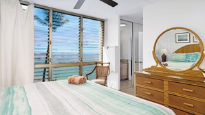 A bedroom with a bed, a dresser, a chair, and a large window with blinds. - Kamaole Nalu 506 - Ocean Front (Kihei)