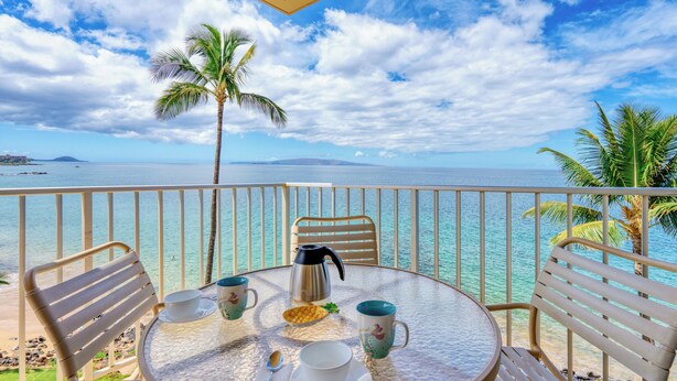 A round table with chairs and a coffee pot, set against a view of the ocean and palm trees. - Kamaole Nalu 506 - Ocean Front (Kihei)