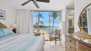 A bedroom with a bed, a chair, a dresser, and a balcony with a view of palm trees and the ocean.