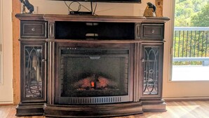 A living room with a fireplace, a television mounted above it, and a large window with a view of a forest.