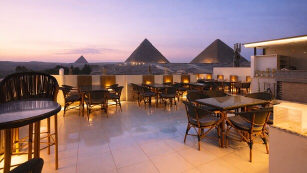 A rooftop terrace with tables and chairs, offering a view of the pyramids at dusk. - Pyramids On The Spot Inn (Giza)