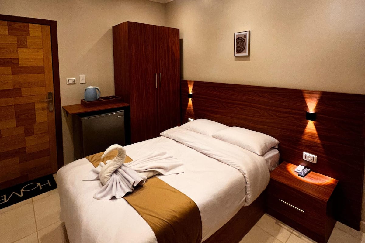 Quarto de hotel com cama, guarda-roupa de madeira, mesa de cabeceira com telefone e porta com placa 'HOME'.