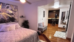 A bedroom with a bed, a ceiling fan, a painting, a small shelf, and a door leading to a kitchen area. - Your private space in an excellent location (Juárez)