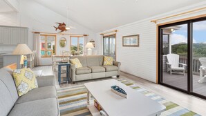 A spacious living area with a sofa, coffee table, and a dining table. There is a ceiling fan, a fish decoration, and a view through a sliding glass door to the outside. - Saltwater Pool, Bar & Ocean Views – Duck OBX (Duck)