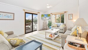 A spacious living room with a large sliding glass door leading to an outdoor deck, a ceiling fan, and a television mounted on the wall. - Saltwater Pool, Bar & Ocean Views – Duck OBX (Duck)