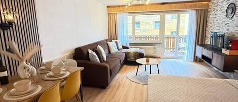 A modern hotel room with a wooden floor, a sofa, a dining table with yellow chairs, a small round table, and a bed.