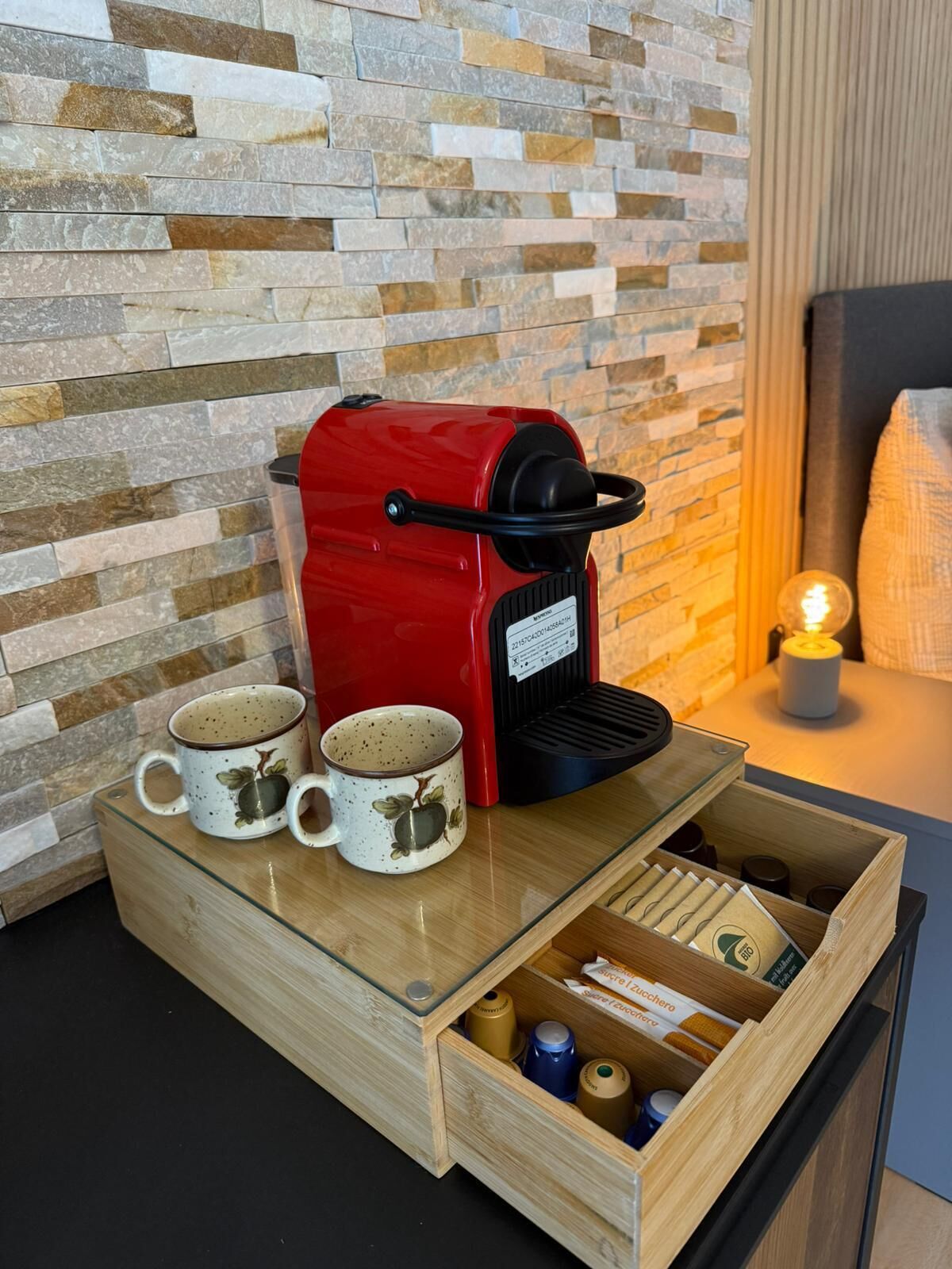 A red coffee machine with two mugs on a wooden tray, a drawer with coffee pods, and a stone wall in the background.