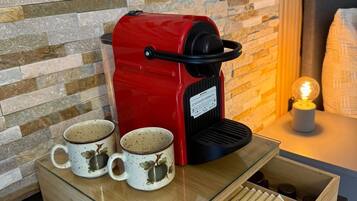 A red coffee machine with two mugs on a wooden tray, a drawer with coffee pods, and a stone wall in the background.