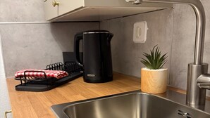 Une cuisine moderne avec un évier en acier inoxydable, une bouilloire noire, un pot de fleurs en bois et un torchon à carreaux rouges et blancs posé sur un étendoir.