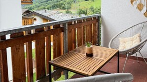 Un balcon avec une table en bois et deux chaises en osier, donnant sur un paysage montagneux.