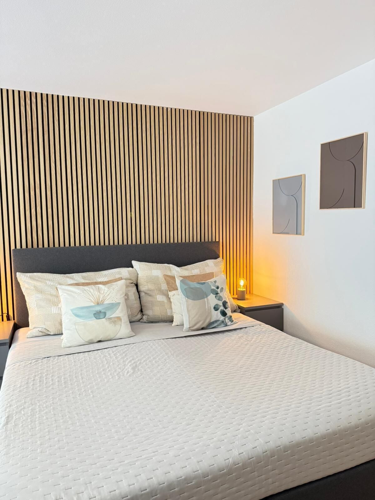 Una cama impecablemente hecha con colcha blanca y varios almohadones, cabecera de listones de madera y dos obras de arte colgadas en la pared.