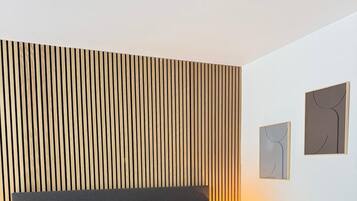 Una cama impecablemente hecha con colcha blanca y varios almohadones, cabecera de listones de madera y dos obras de arte colgadas en la pared.