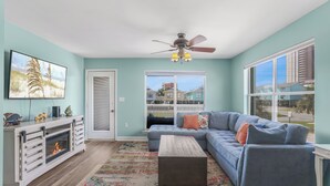 A living room with a sectional sofa, a fireplace, and a large window with a view of buildings. - Beach Days & Fire Nights! Family 3BR Duplex Just Steps from the Sand! (Gulf Shores)
