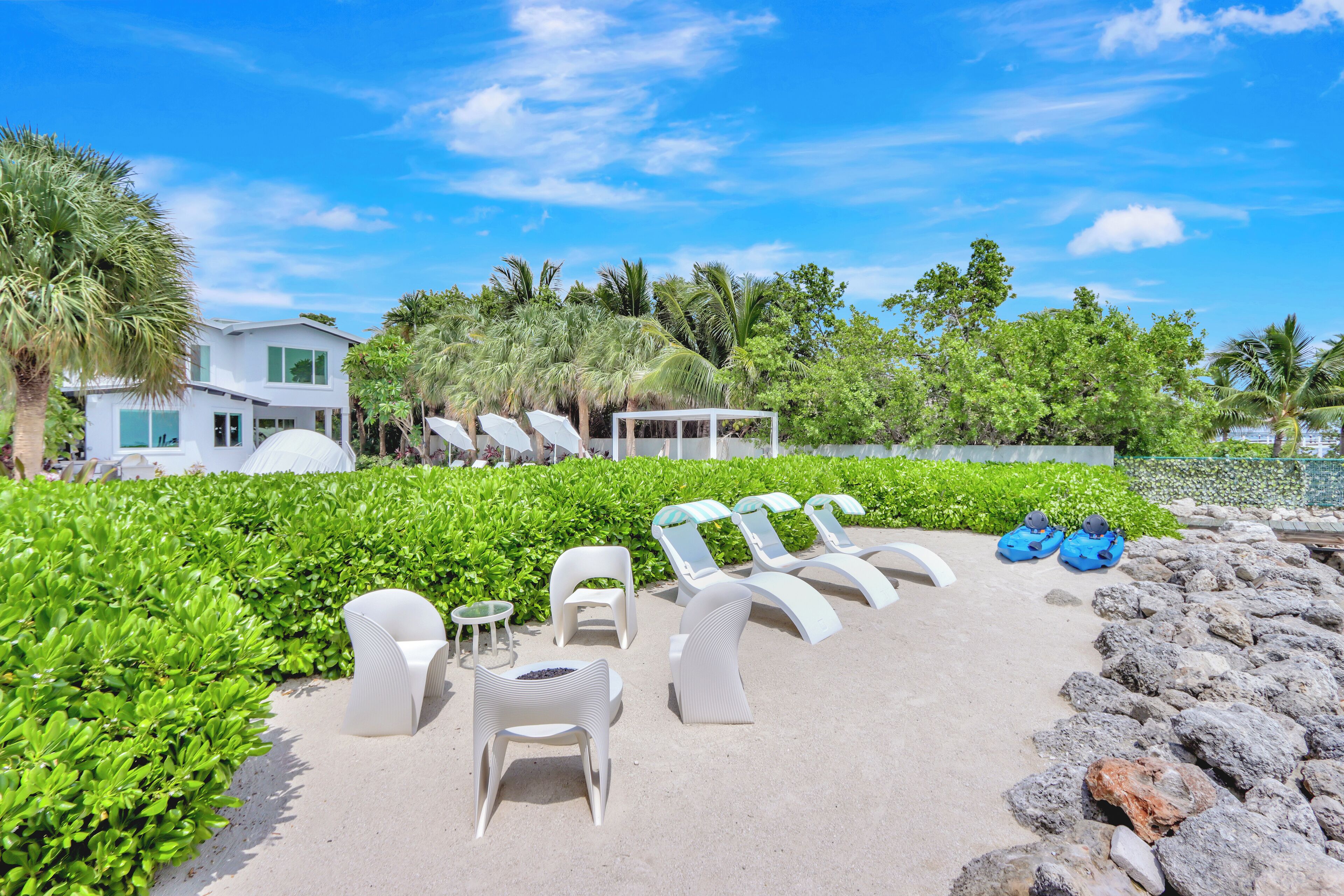 A beachside lounge area with white lounge chairs and a small table, surrounded by lush greenery and a clear blue sky.