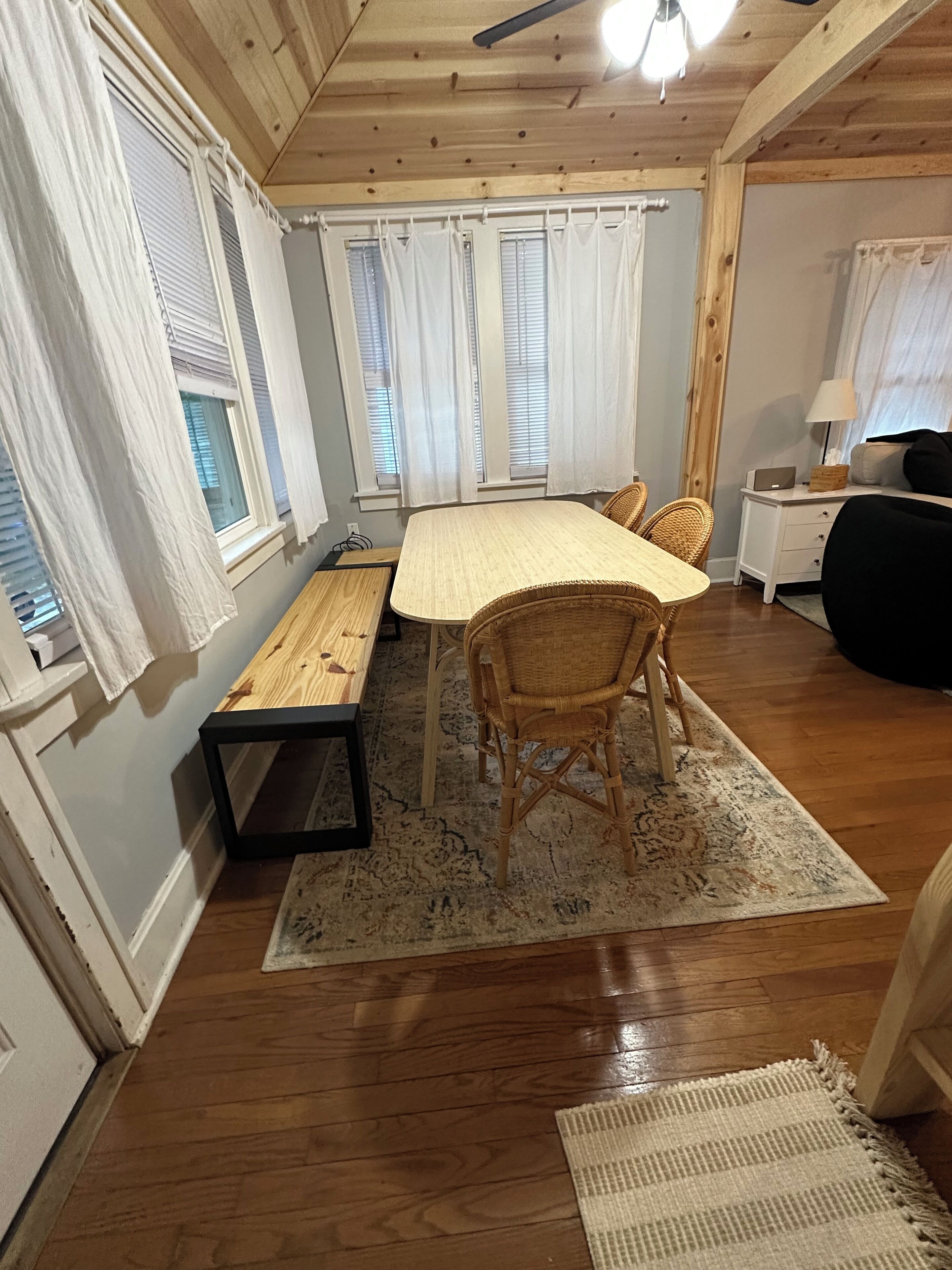 A cozy room with a wooden ceiling, a dining table with chairs, a bench, and a small white nightstand with a lamp.