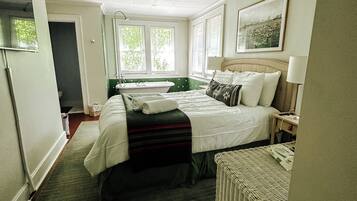 Habitación de hotel con cama, ventilador de techo, ventana con persianas, un cuadro en la pared y una mesita con lámpara.
