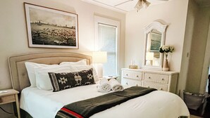 A neatly made bed with a green bedspread, a white blanket, and a decorative pillow. There is a nightstand with a lamp and a framed picture on the wall.