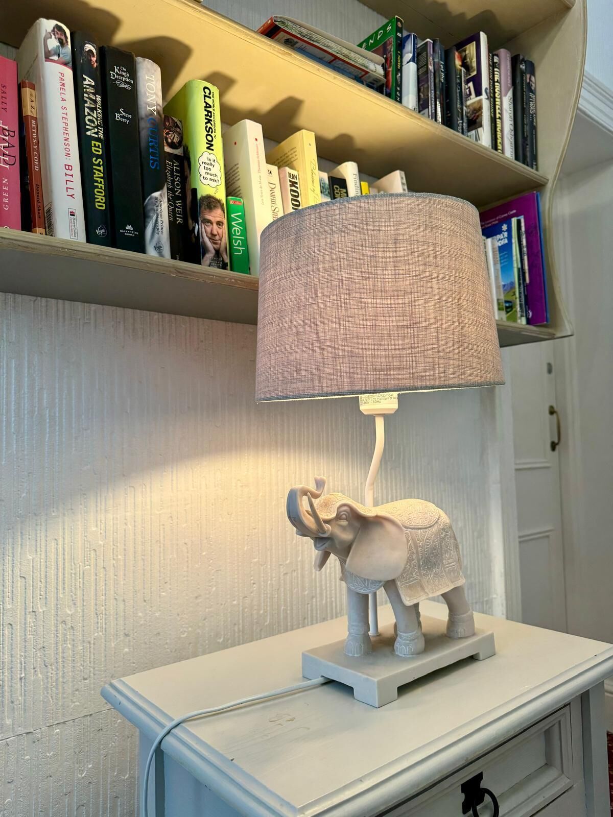 Una lámpara con base de elefante sobre una mesa blanca, una estantería con varios libros y una pared blanca al fondo.