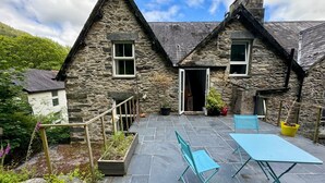 A stone house with a steeply pitched roof, a small porch with a wooden railing, and a door leading to a covered entrance. - Fairhaven Stay (Betws-Y-Coed)
