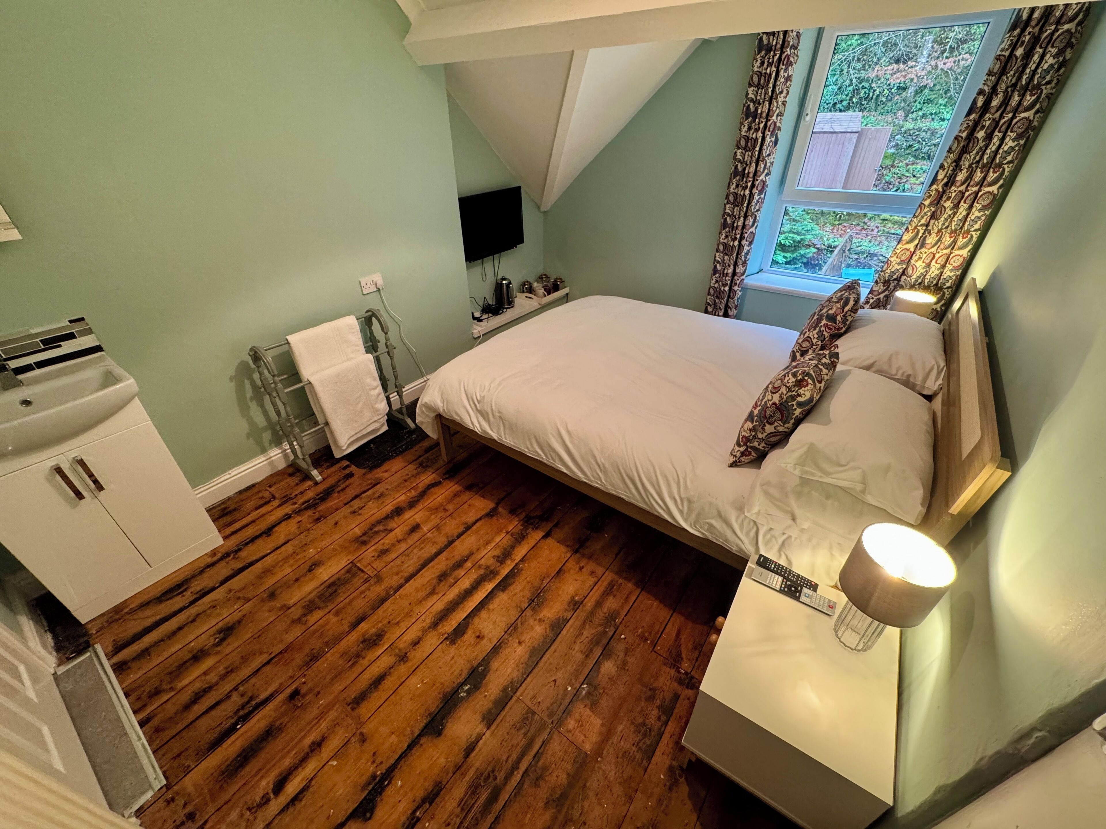 A bedroom with a wooden floor, a bed with white bedding, a nightstand, a TV, and a window with curtains.