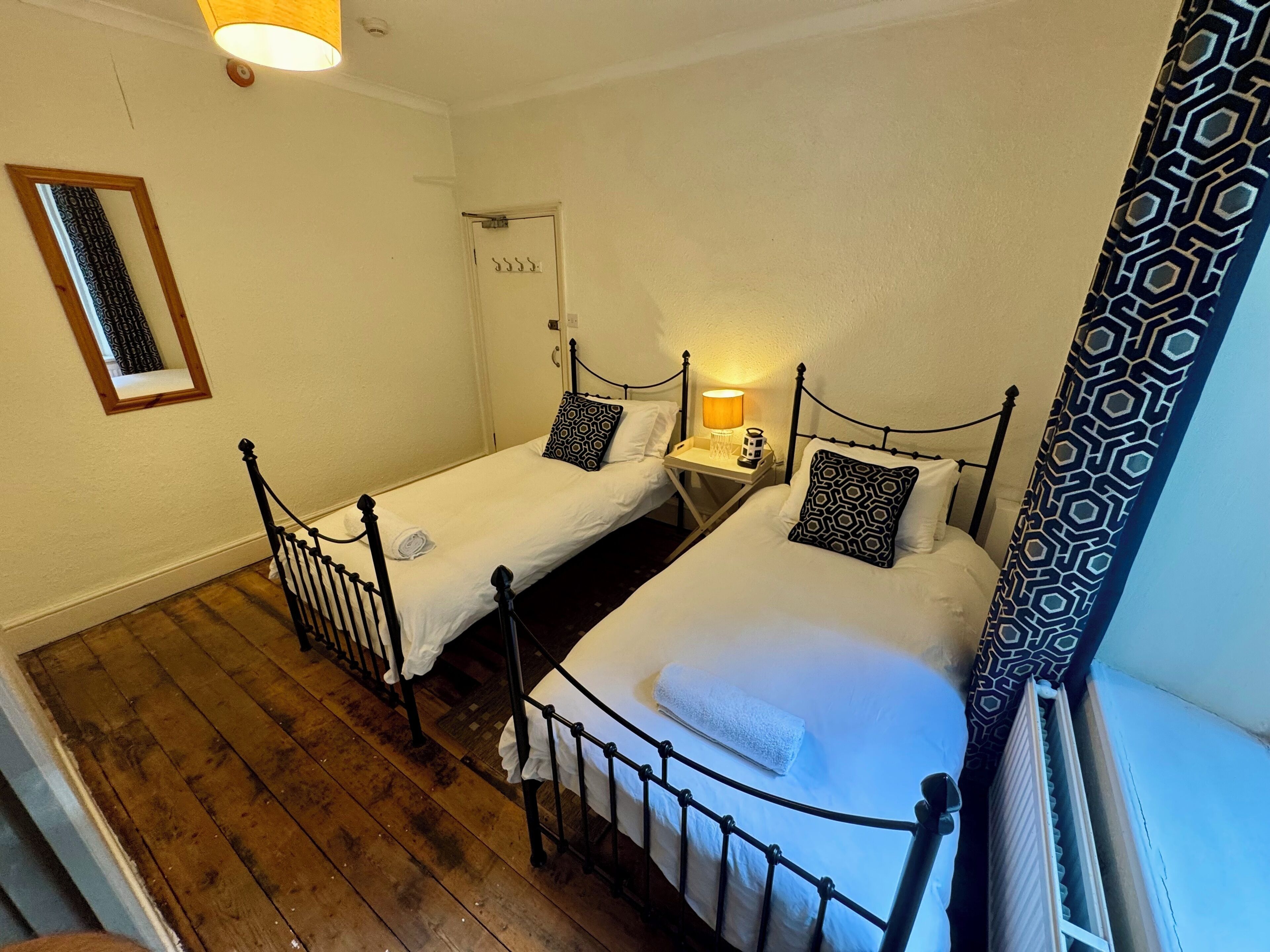 Um quarto pequeno e acolhedor com duas camas de solteiro, piso de madeira, uma mesinha com abajur e um espelho na parede.