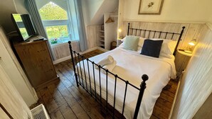 A bedroom with a wooden floor, a metal bed frame, a white bedspread, a bedside table with a lamp, and a window with curtains. - Fairhaven Stay (Betws-Y-Coed)