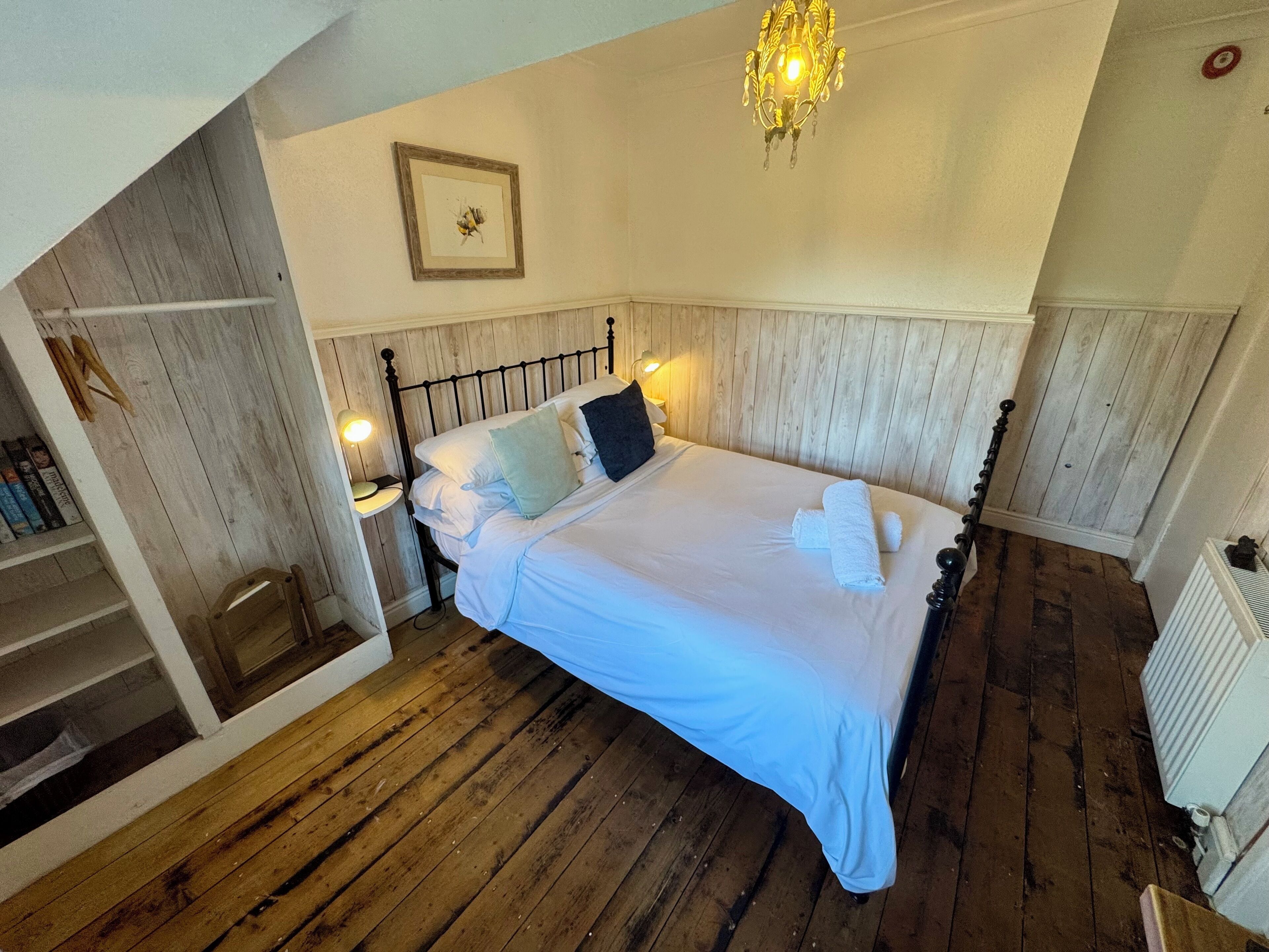 A bedroom with a wooden floor, a bed with white linens, a nightstand with a lamp, and a chandelier.