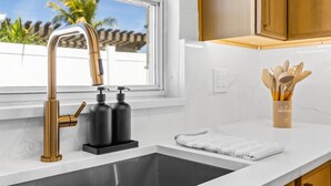A kitchen with a stainless steel sink, a gold faucet, and a window with a view of palm trees.