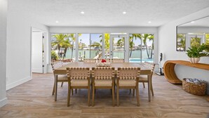 A spacious dining area with a large table and chairs, a view of a pool and palm trees, and a modern curved console table with a plant.