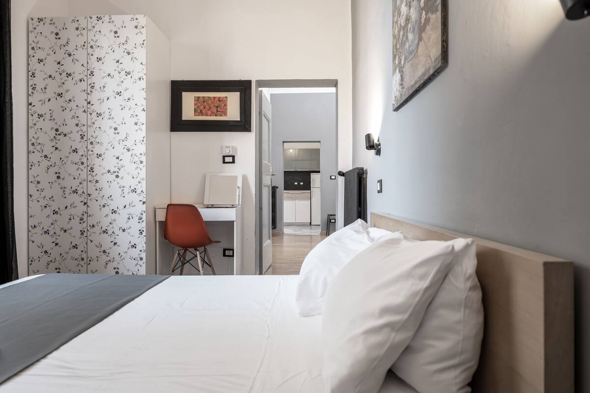 A hotel room with a bed, a desk, a chair, a framed picture on the wall, and a view into the kitchen area.
