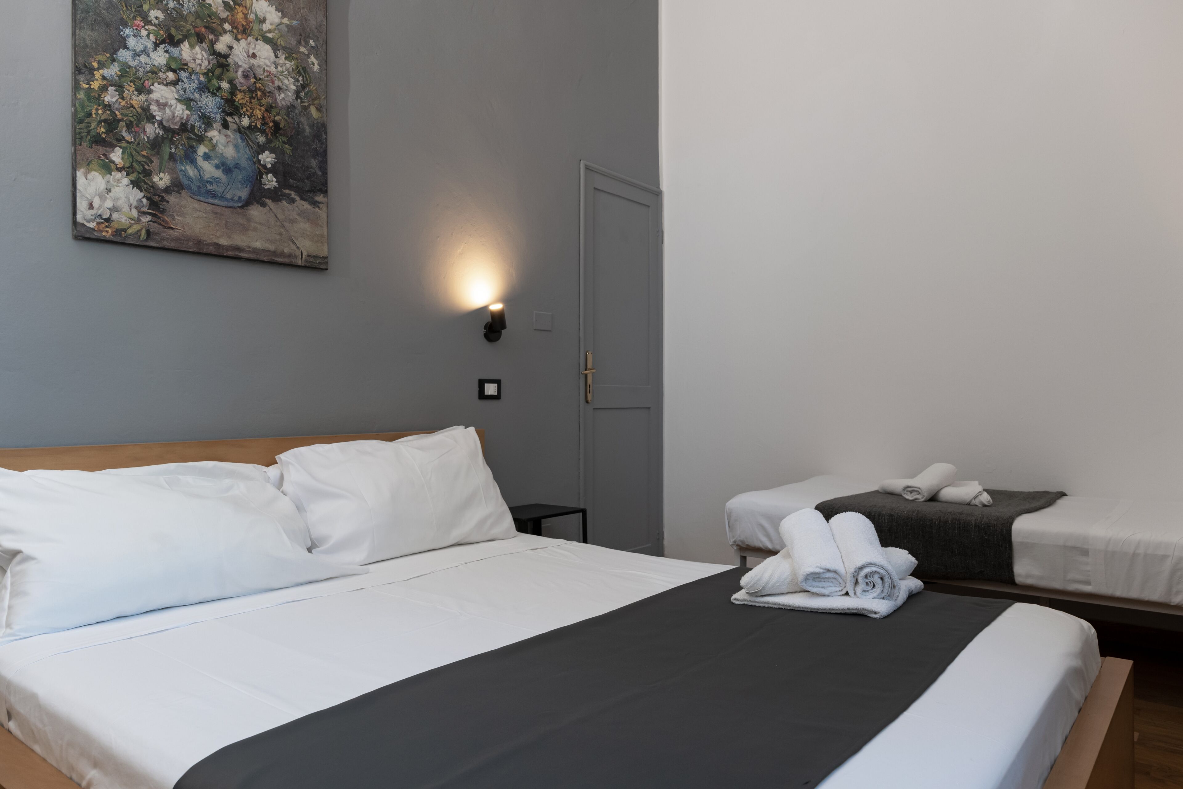Habitación de hotel con una cama grande, una cama más pequeña, una mesita de noche, una lámpara de pared y un cuadro.