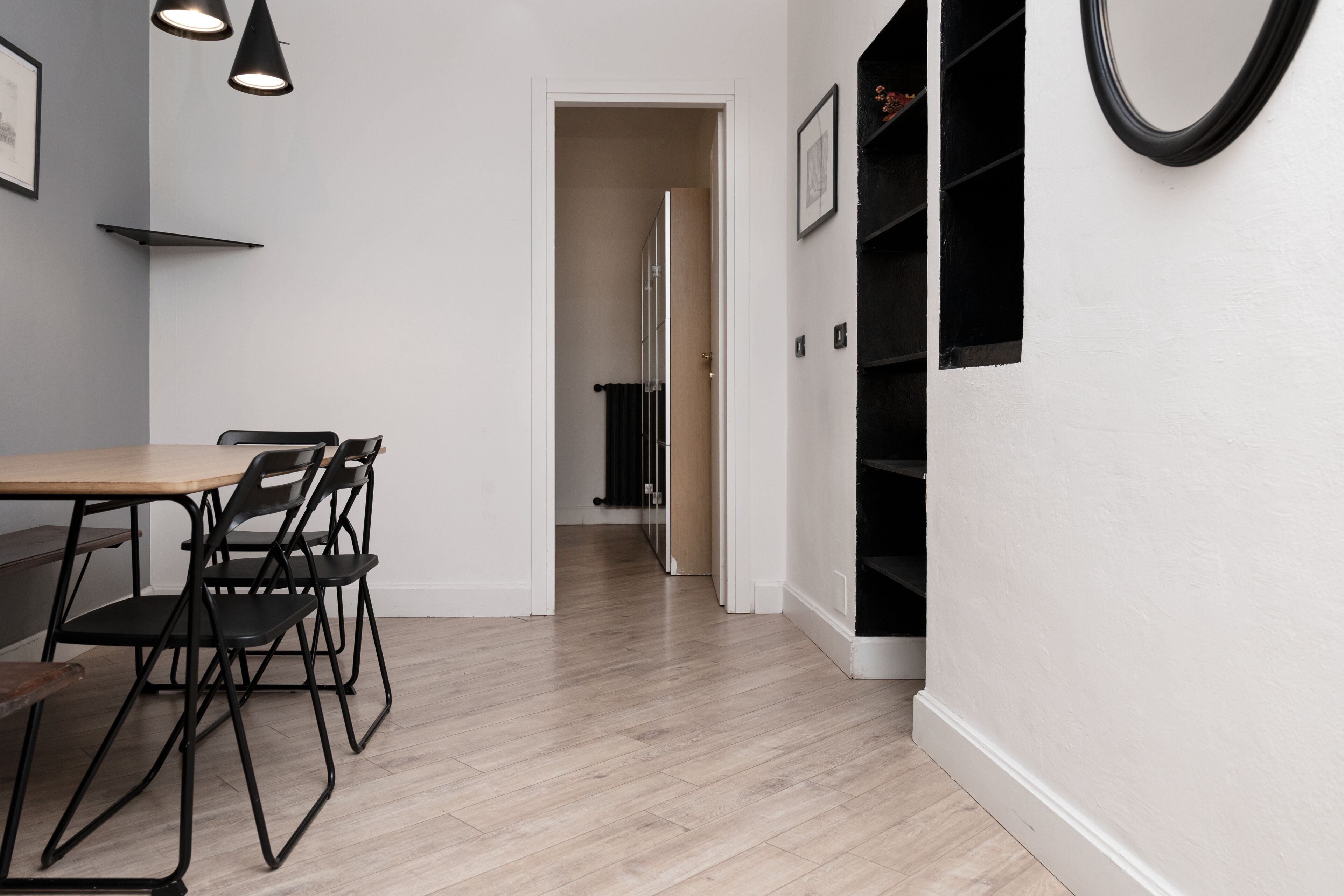 A modern interior with a wooden dining table, black chairs, a white wall, and a hallway leading to a room with a black door.