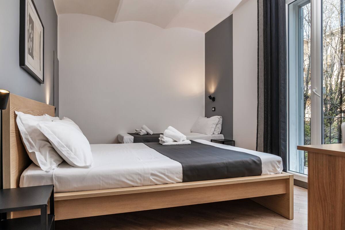 A modern bedroom with a single bed, bedside tables, a nightstand, and a large window with curtains.
