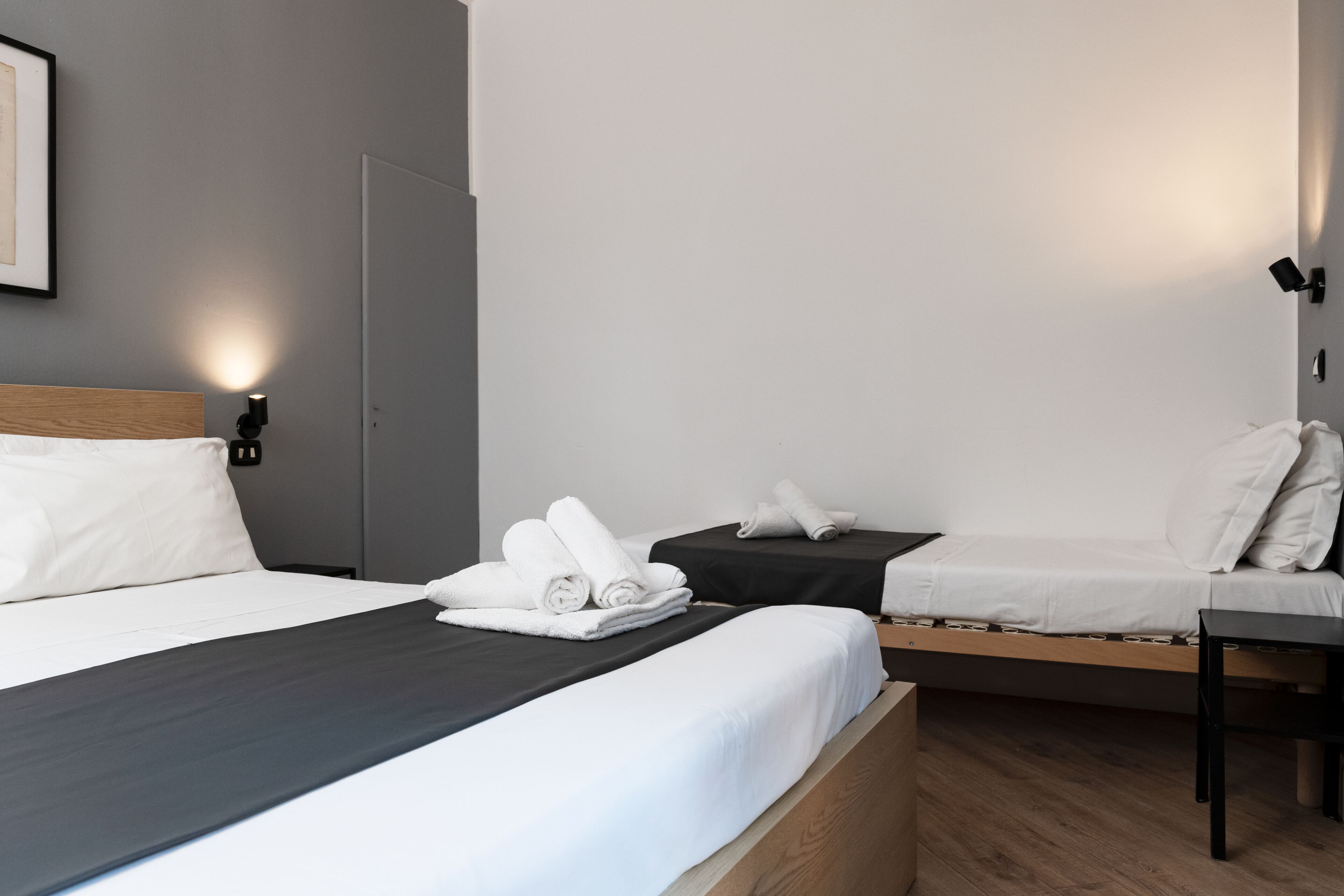 Habitación de hotel con una cama grande, una cama más pequeña, un escritorio y una lámpara de pared.
