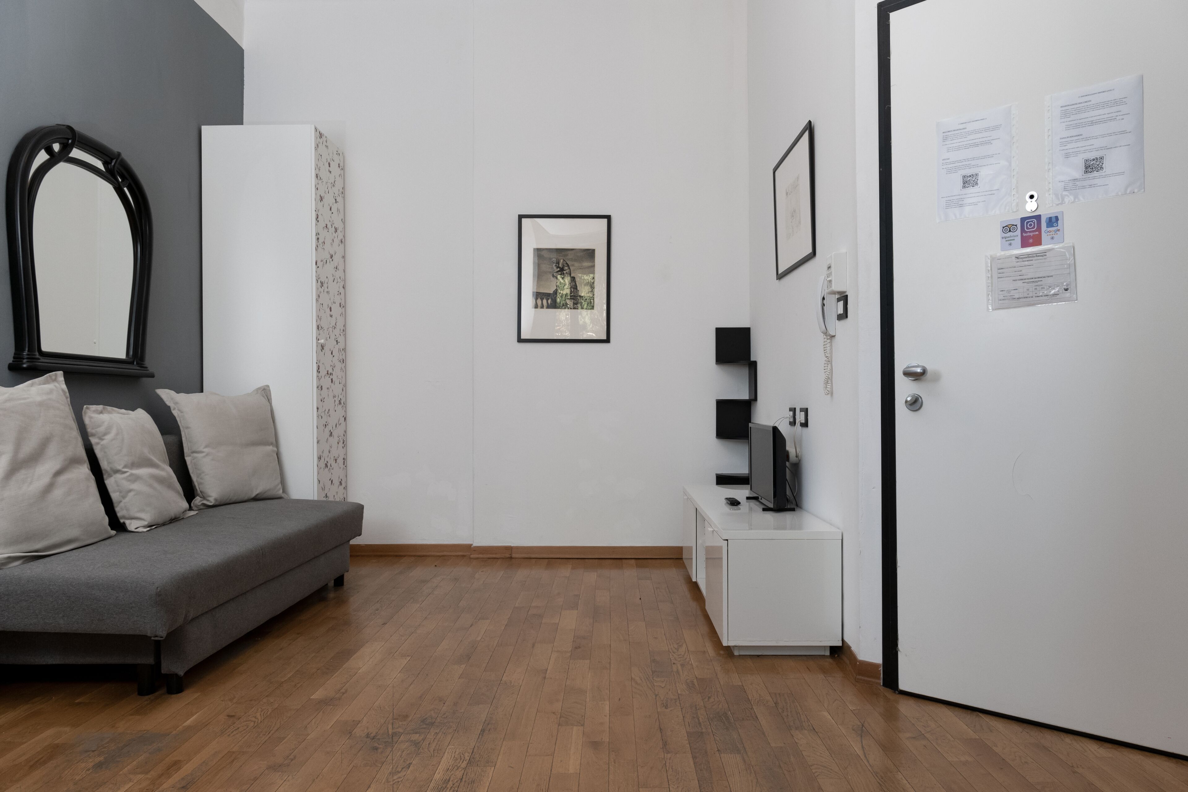 Ein Zimmer mit grauem Sofa, einem weißen Schrank, einem gerahmten Bild an der Wand, einem Fernseher auf einem kleinen Tisch und einer Tür mit angebrachten Papieren.