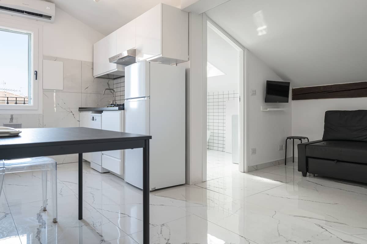 A modern kitchen with white cabinets, a black countertop, and a black sofa in a bright room with a tiled floor.