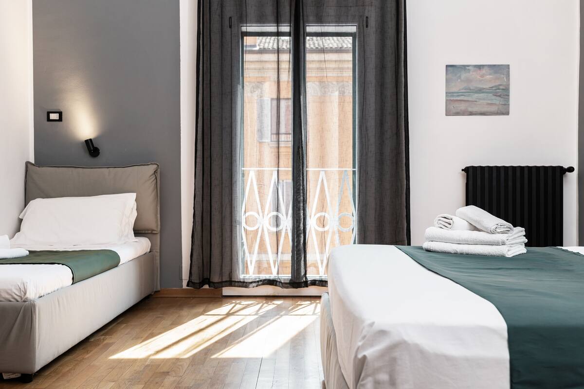 A hotel room with two beds, a view of the outside, a painting on the wall, and a black radiator.