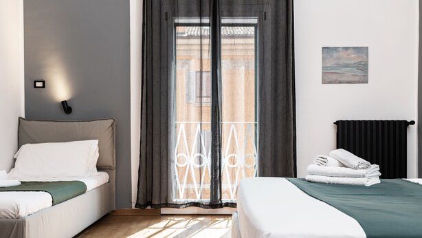 A hotel room with two beds, a view of the outside, a painting on the wall, and a black radiator. - Saragozza Apartments (Bologna)